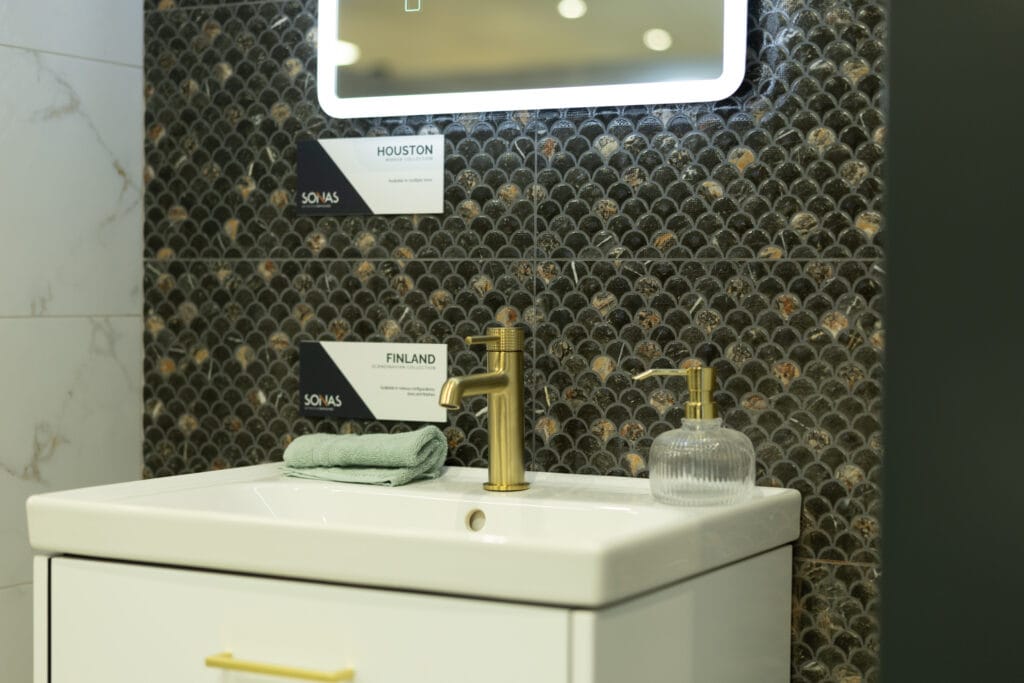 Brushed gold basin tap on a white vanity unit in front of dark fish-scale mosaic tiles